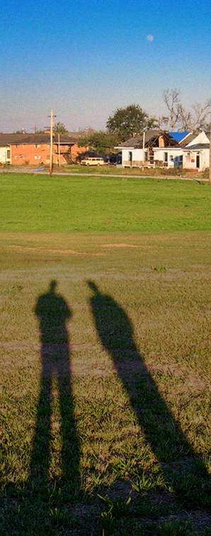 A photograph showing the shadows of Post-K DuMarais and Timezone LaFontaine standing on a levee in New Orleans. In the distant background are homes damaged by Hurricane Katrina.